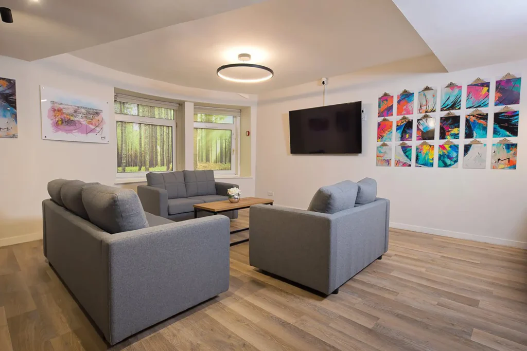 A shared living space in student accommodation. Three two-seater sofas around a table. There is a large TV on the wall. The room is well lit.