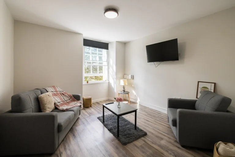 A shared living space at Lyon Street. The room is spacious and light, with a two person sofa and a single comfy chair. There is a TV attached to a wall. In the middle of the room is a coffee table